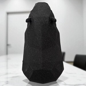 May include: A black, geometric-shaped object with a textured surface. The object has a tall, tapered form with a flat base and two small, ear-like protrusions near the top. It is set on a white marble-like surface.