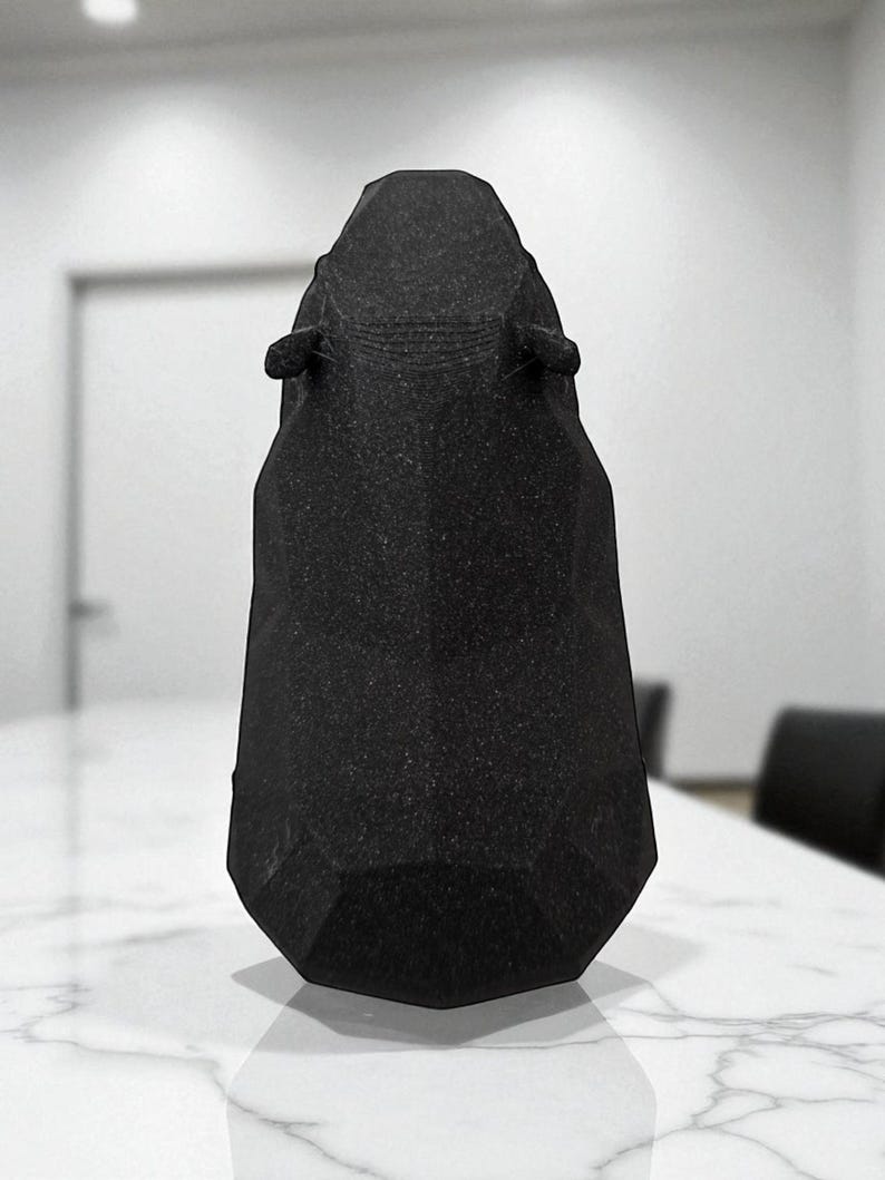 May include: A black, geometric-shaped object with a textured surface. The object has a tall, tapered form with a flat base and two small, ear-like protrusions near the top. It is set on a white marble-like surface.