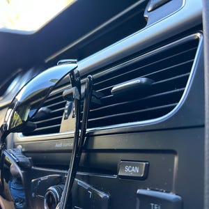 May include: A pair of black sunglasses and a black hair clip are hanging from the air vent in a car. The car's dashboard is visible, with buttons labeled "SCAN" and "TEL".