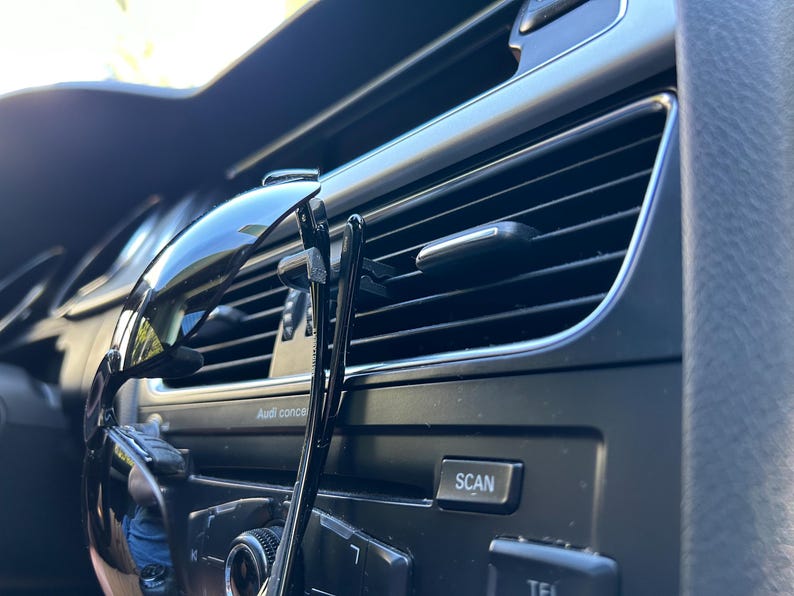 May include: A pair of black sunglasses and a black hair clip are hanging from the air vent in a car. The car's dashboard is visible, with buttons labeled "SCAN" and "TEL".