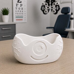 May include: A white, eye-shaped bowl with a smiling face design. The bowl has a textured surface and is likely made of ceramic or a similar material. The design includes an eye with a circular pupil and vein-like details.
