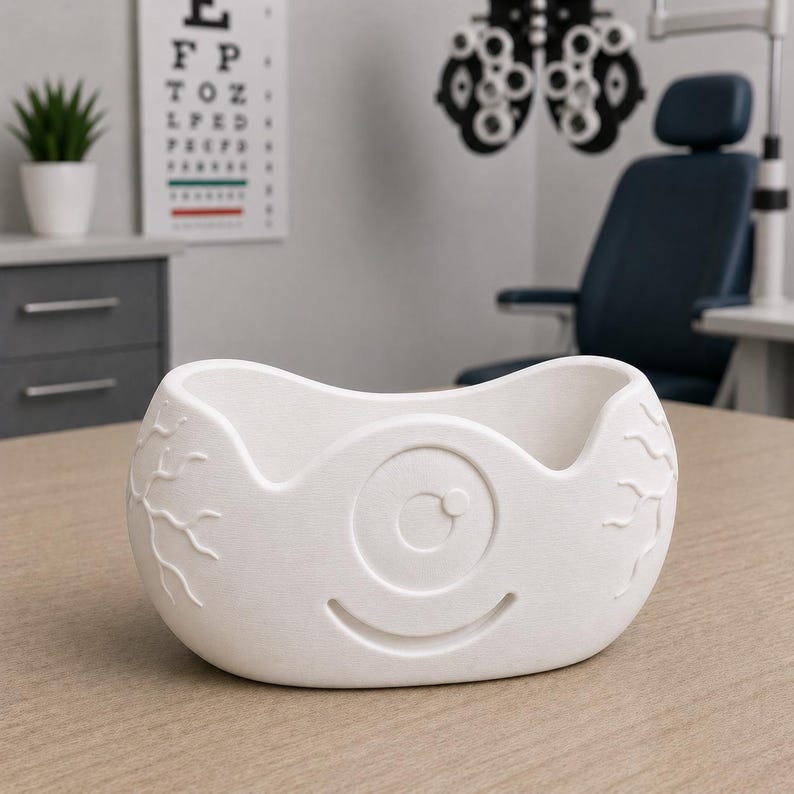 May include: A white, eye-shaped bowl with a smiling face design. The bowl has a textured surface and is likely made of ceramic or a similar material. The design includes an eye with a circular pupil and vein-like details.