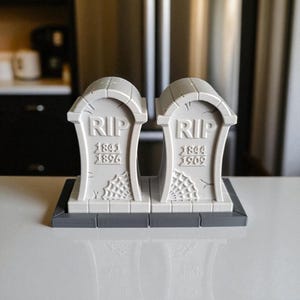 May include: Two gray, tombstone-shaped decorations with the letters "RIP" at the top. Each tombstone has dates and a spiderweb design. The tombstones are on a gray base, perfect for Halloween or spooky decor.