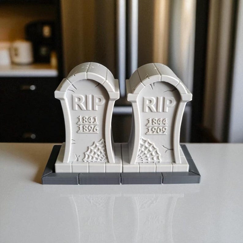 May include: Two gray, tombstone-shaped decorations with the letters "RIP" at the top. Each tombstone has dates and a spiderweb design. The tombstones are on a gray base, perfect for Halloween or spooky decor.