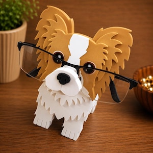 May include: A novelty eyeglass holder in the shape of a dog. The holder is white, tan, and black, with a black nose and black-framed glasses. The holder is on a wooden surface.