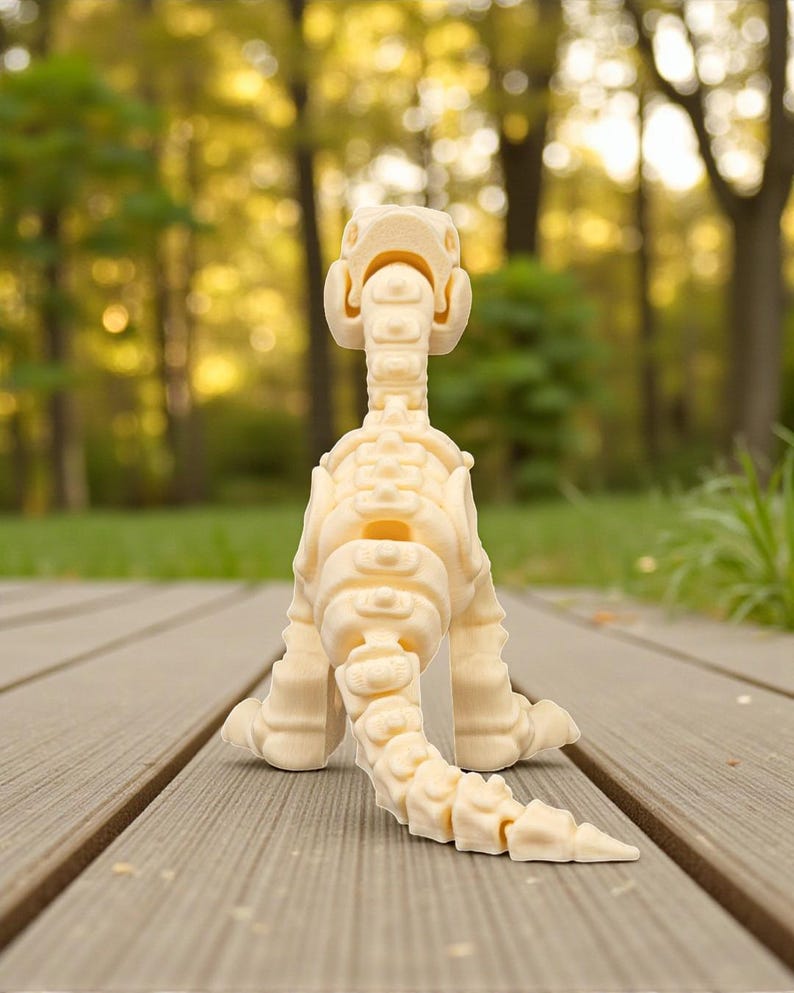 May include: A cream-colored dinosaur skeleton figurine, posed on a wooden deck. The toy dinosaur is facing away from the camera, showcasing its spine, tail, and leg structure. The background is a blurred view of a green lawn and trees.