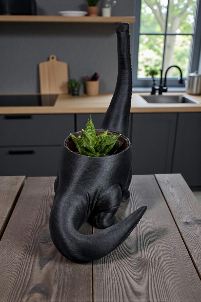 May include: A black dinosaur-shaped plant pot containing a green succulent. The pot is made of a textured material and is on a wooden table. The background shows a kitchen with dark cabinets and a window.