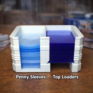 Puede incluir: Un soporte de tarjetas de pl&aacute;stico blanco con dos compartimentos. Un lado contiene una pila de fundas protectoras azules, y el otro una pila de top loaders azules. El texto "Penny Sleeves" y "Top Loaders" est&aacute; impreso en la base.