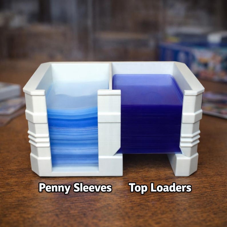 Puede incluir: Un soporte de tarjetas de pl&aacute;stico blanco con dos compartimentos. Un lado contiene una pila de fundas protectoras azules, y el otro una pila de top loaders azules. El texto "Penny Sleeves" y "Top Loaders" est&aacute; impreso en la base.
