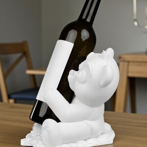 May include: A white bear-shaped wine bottle holder. The bear is holding a dark glass wine bottle with a blank white label. The holder is sitting on a wooden table. The background includes a chair and a lamp.