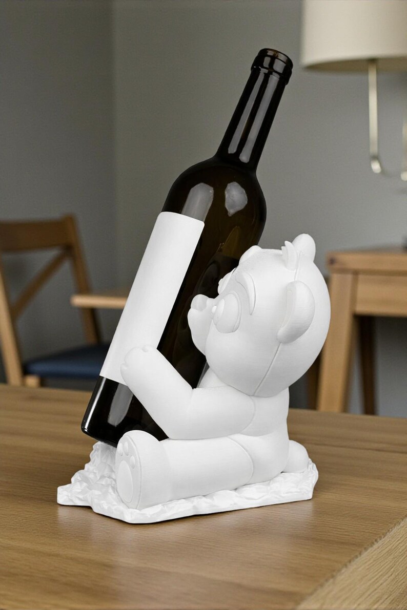 May include: A white bear-shaped wine bottle holder. The bear is holding a dark glass wine bottle with a blank white label. The holder is sitting on a wooden table. The background includes a chair and a lamp.