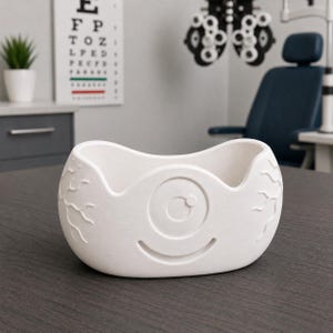 May include: A white, eye-shaped decorative bowl with a smiling face design. The bowl features an embossed eye and vein details, perfect for an optometrist's office or as a unique home decor item.