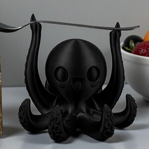 Puede incluir: Un soporte para utensilios negro con forma de pulpo con una cuchara equilibrada sobre sus tent&aacute;culos levantados. El soporte tiene un dise&ntilde;o de dibujos animados con ojos grandes y una boca sonriente. Un cuenco de fruta est&aacute; en el fondo.