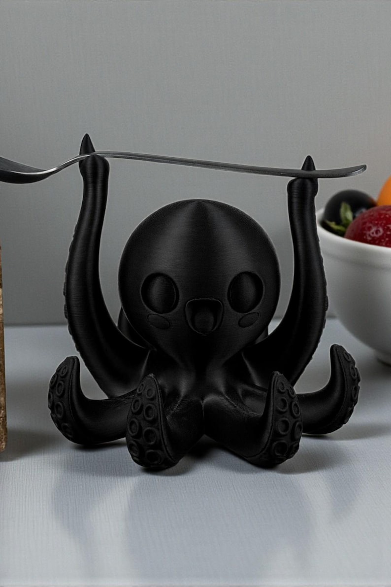 Puede incluir: Un soporte para utensilios negro con forma de pulpo con una cuchara equilibrada sobre sus tent&aacute;culos levantados. El soporte tiene un dise&ntilde;o de dibujos animados con ojos grandes y una boca sonriente. Un cuenco de fruta est&aacute; en el fondo.