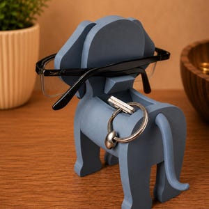 May include: A blue elephant-shaped desk organizer holding black-framed glasses and a silver key ring. The organizer is made of wood and has a slot for keys and a top for glasses. The elephant has a trunk, ears, and a tail.