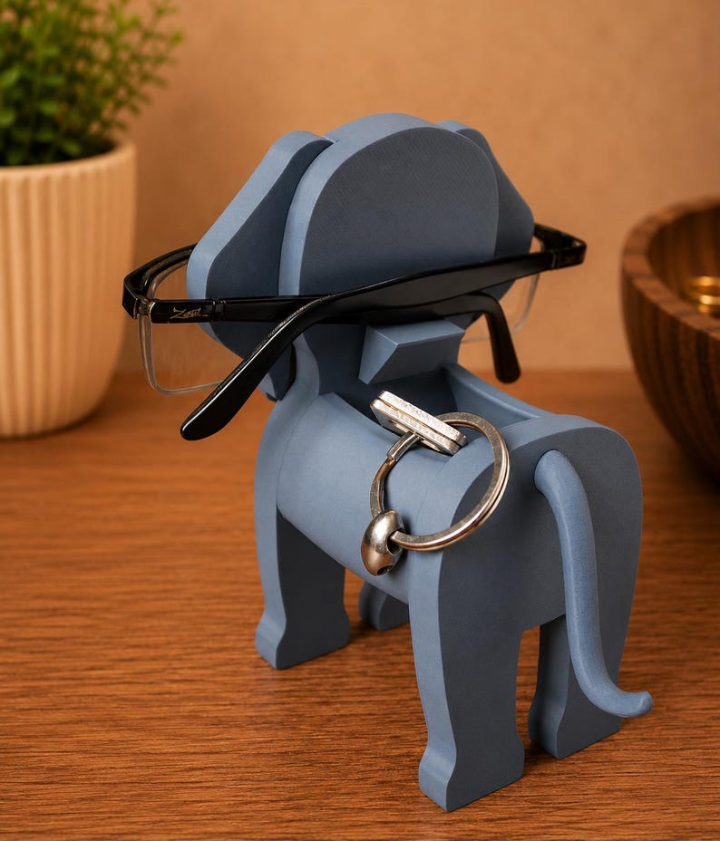 May include: A blue elephant-shaped desk organizer holding black-framed glasses and a silver key ring. The organizer is made of wood and has a slot for keys and a top for glasses. The elephant has a trunk, ears, and a tail.