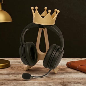 May include: A wooden headphone stand with a gold crown on top, holding black headphones with a microphone. The stand is on a wooden surface, with a gold lamp and a brown leather notebook in the background.