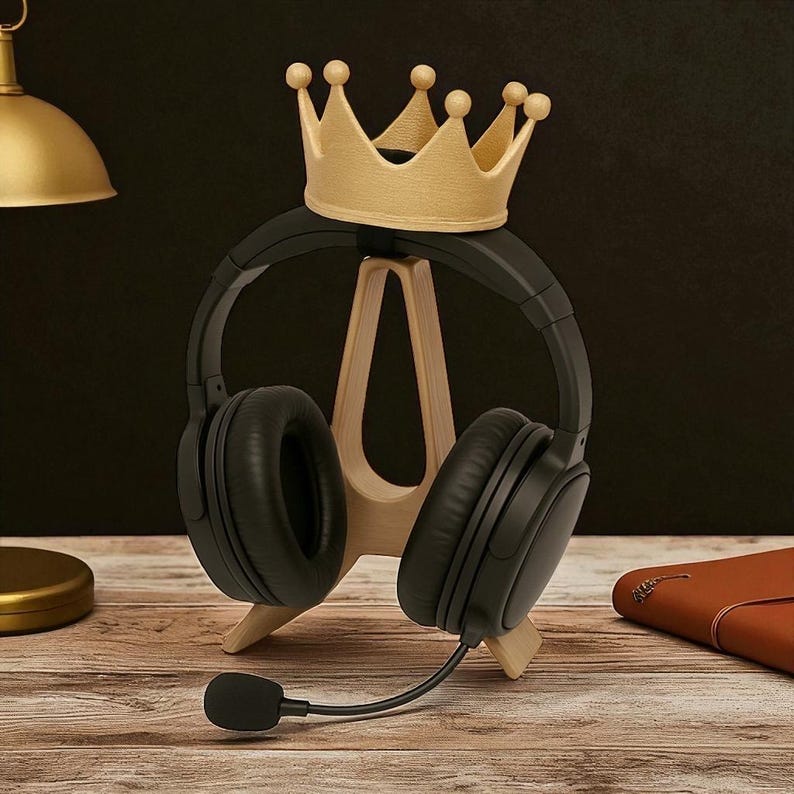 May include: A wooden headphone stand with a gold crown on top, holding black headphones with a microphone. The stand is on a wooden surface, with a gold lamp and a brown leather notebook in the background.
