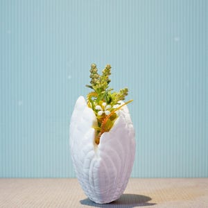 May include: A white, textured vase shaped like a swan's wings, holding a small arrangement of artificial flowers. The flowers include white and yellow blossoms with green and orange stems. The background is a light blue wall with vertical stripes.
