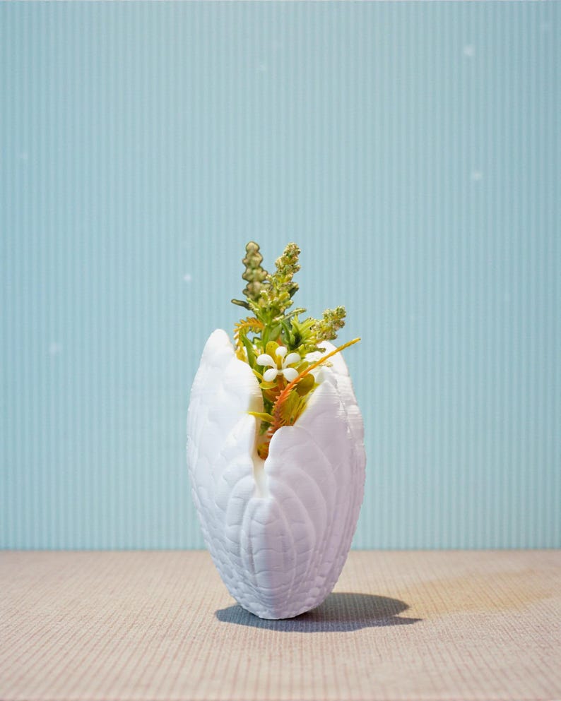 May include: A white, textured vase shaped like a swan's wings, holding a small arrangement of artificial flowers. The flowers include white and yellow blossoms with green and orange stems. The background is a light blue wall with vertical stripes.