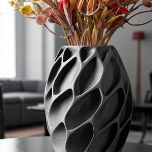 May include: A dark gray, textured vase with an open, geometric design, holding colorful artificial flowers. The vase has an oval shape and sits on a dark surface. The flowers are a mix of yellow, orange, and red.