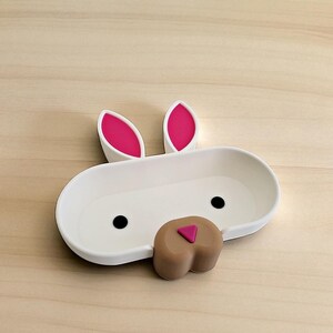 May include: A white bunny-shaped dish with pink-lined ears, black eyes, and a brown nose with a pink triangle. The dish is oval-shaped and sits on a light wood surface. A decorative item for the home.