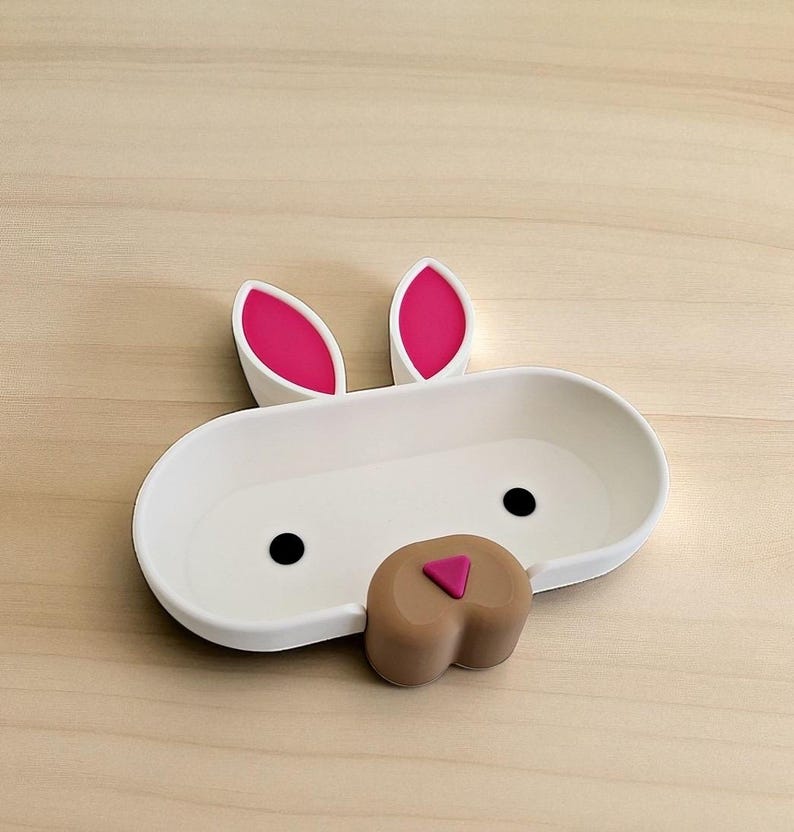 May include: A white bunny-shaped dish with pink-lined ears, black eyes, and a brown nose with a pink triangle. The dish is oval-shaped and sits on a light wood surface. A decorative item for the home.