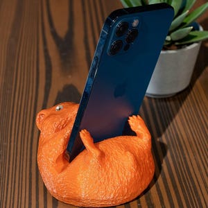 K&ouml;nnte beinhalten: Ein orangefarbener Telefonst&auml;nder in Form eines liegenden Hamsters, der ein blaues Smartphone h&auml;lt. Der St&auml;nder hat eine strukturierte Oberfl&auml;che und steht auf einem Holztisch. Im Hintergrund ist eine kleine Topfpflanze zu sehen.