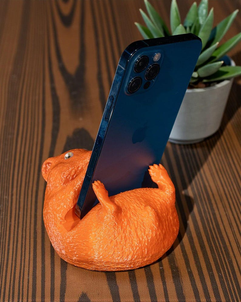 K&ouml;nnte beinhalten: Ein orangefarbener Telefonst&auml;nder in Form eines liegenden Hamsters, der ein blaues Smartphone h&auml;lt. Der St&auml;nder hat eine strukturierte Oberfl&auml;che und steht auf einem Holztisch. Im Hintergrund ist eine kleine Topfpflanze zu sehen.