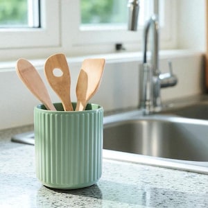 May include: A kitchen setting with a light green utensil holder containing wooden spoons and spatulas. A glass jar with a wooden lid and a stainless steel sink are also visible. The scene is bright and airy, suggesting a modern kitchen.