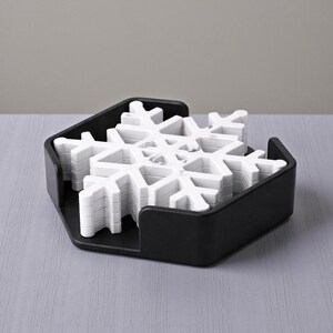Puede incluir: Un juego de posavasos blancos con forma de copo de nieve en un soporte hexagonal negro. Los posavasos est&aacute;n apilados y listos para usar. El soporte tiene un dise&ntilde;o sencillo y moderno, ideal para bebidas.