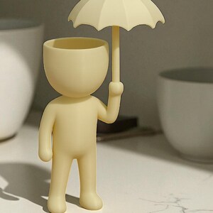 May include: A cream-colored figurine of a person holding an umbrella. The figurine has a rounded head and body with a small opening at the top. The umbrella is also cream-colored and has a curved shape.