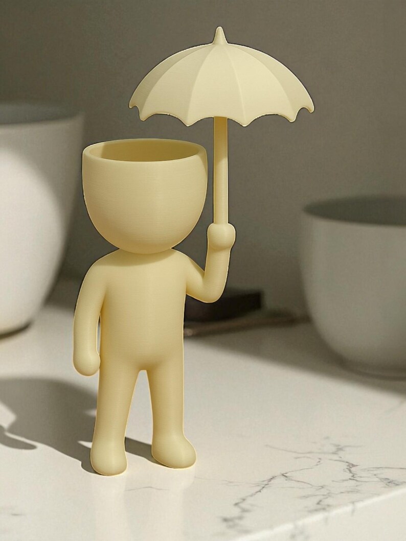 May include: A cream-colored figurine of a person holding an umbrella. The figurine has a rounded head and body with a small opening at the top. The umbrella is also cream-colored and has a curved shape.