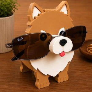 May include: A brown and white dog-shaped sunglasses holder with black sunglasses. The holder has a white snout, black eyes, and a red tongue. The sunglasses have a Ray-Ban logo.