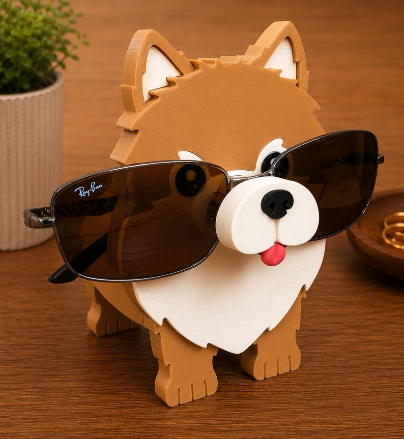 May include: A brown and white dog-shaped sunglasses holder with black sunglasses. The holder has a white snout, black eyes, and a red tongue. The sunglasses have a Ray-Ban logo.
