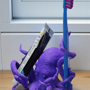 Puede incluir: Un soporte para cepillo de dientes con forma de pulpo morado con un cepillo de dientes azul y un tubo de pasta de dientes. El cepillo de dientes tiene un mango azul y cerdas rosas. El tubo de pasta de dientes es negro y blanco.