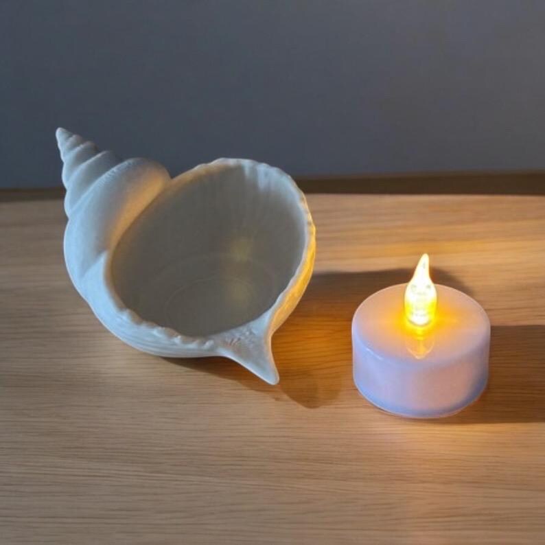 May include: A white ceramic seashell-shaped bowl and a small, round, white LED candle with a flickering yellow light. The candle is placed on a wooden surface, creating a warm, inviting ambiance. The seashell bowl adds a touch of coastal decor.