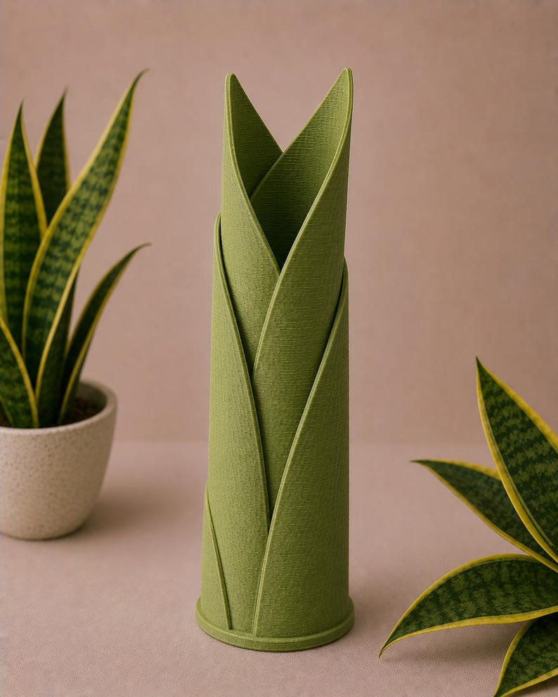 May include: A green, cylindrical vase with a leaf-like design. The vase is made of a textured material and has a round base. Two potted snake plants with yellow-edged green leaves are in the background.