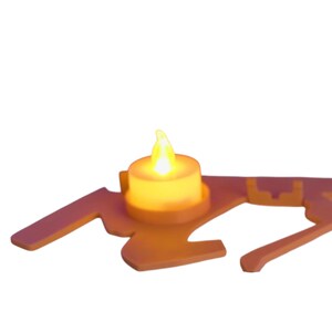 Puede incluir: Una peque&ntilde;a vela que funciona con pilas con una llama amarilla parpadeante se encuentra sobre un soporte decorativo de color naranja claro. El soporte tiene una forma estilizada &uacute;nica con brazos extendidos y una base circular para la vela.