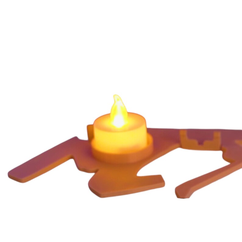 Puede incluir: Una peque&ntilde;a vela que funciona con pilas con una llama amarilla parpadeante se encuentra sobre un soporte decorativo de color naranja claro. El soporte tiene una forma estilizada &uacute;nica con brazos extendidos y una base circular para la vela.