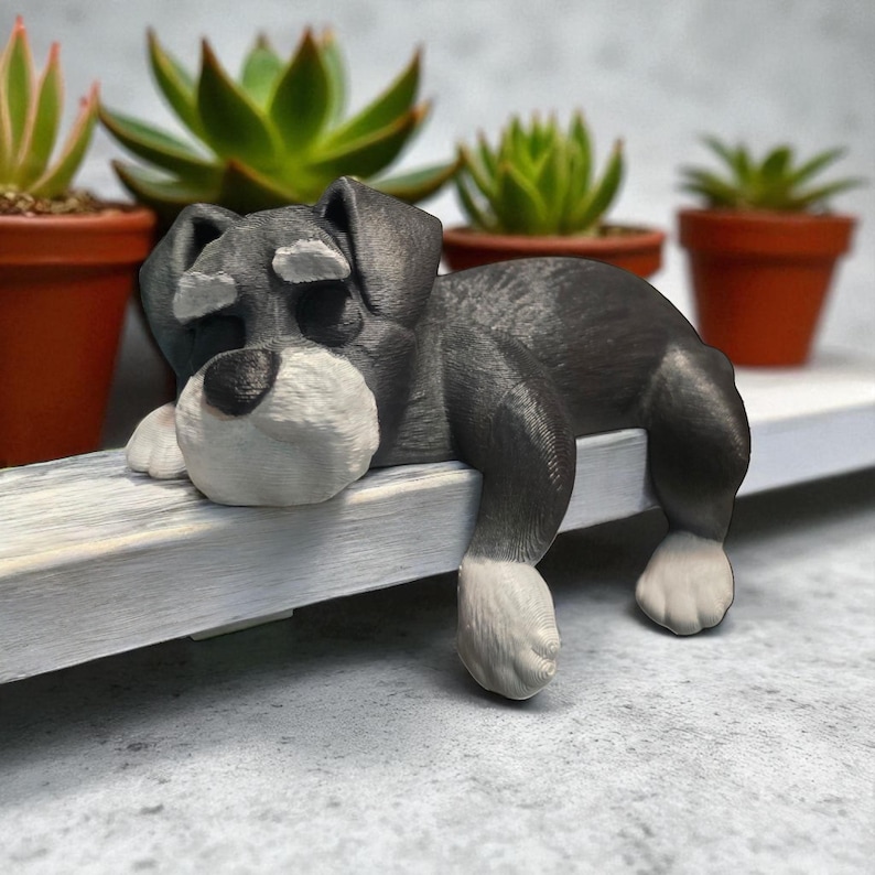 May include: A gray and white figurine of a dog, resting on a white ledge. The dog has a black body, white paws, and a white muzzle. Behind the dog are several small potted succulents.