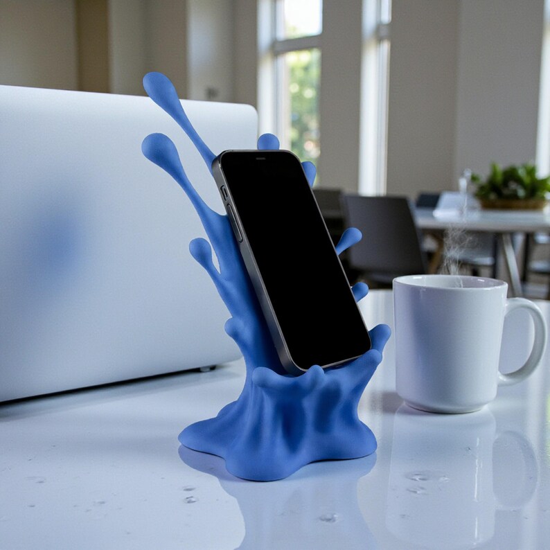 May include: A blue phone stand shaped like a splash, holding a black smartphone. The stand is on a white surface, with a laptop and a white mug in the background. The stand's design features fluid, dynamic lines.