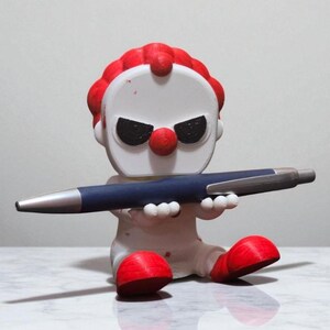 May include: A white and red clown figurine with a red afro, nose, and feet, holding a blue pen with a silver tip. The clown has black eye markings and is sitting down. The pen is held horizontally.