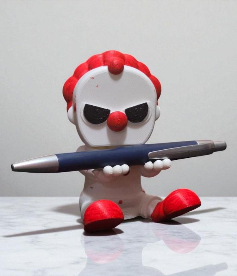 May include: A white and red clown figurine with a red afro, nose, and feet, holding a blue pen with a silver tip. The clown has black eye markings and is sitting down. The pen is held horizontally.