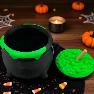 May include: A black cauldron with a green rim and a skull design, next to a green, textured lid with a wooden stick. The cauldron sits on a black lace doily, surrounded by candy corn and miniature orange pumpkins, evoking a Halloween theme.