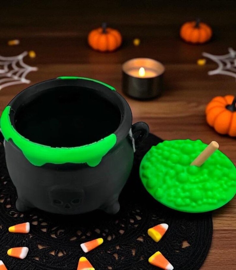 May include: A black cauldron with a green rim and a skull design, next to a green, textured lid with a wooden stick. The cauldron sits on a black lace doily, surrounded by candy corn and miniature orange pumpkins, evoking a Halloween theme.