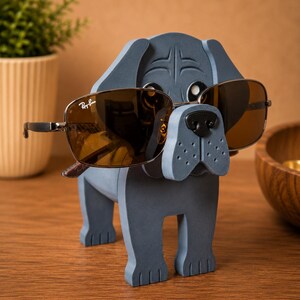 May include: A gray wooden dog-shaped eyeglass holder with brown sunglasses. The sunglasses have a metal frame and the Ray-Ban logo. The dog has a black nose and is standing on a wooden surface.
