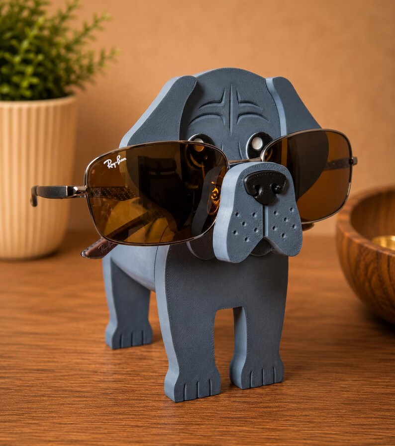 May include: A gray wooden dog-shaped eyeglass holder with brown sunglasses. The sunglasses have a metal frame and the Ray-Ban logo. The dog has a black nose and is standing on a wooden surface.