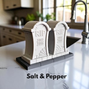 May include: A gray salt and pepper shaker set shaped like tombstones, with the text "RIP" and dates. Each tombstone has a spiderweb design. The base is dark gray, and the text "Salt & Pepper" is below the set.