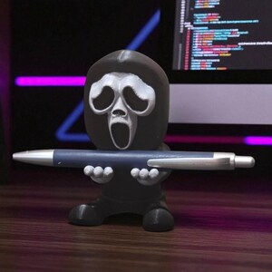 Puede incluir: Un portal&aacute;pices negro con forma de m&aacute;scara de Ghostface de las pel&iacute;culas Scream, sosteniendo un bol&iacute;grafo azul. El portal&aacute;pices tiene una cara blanca y est&aacute; sobre una superficie de madera.
